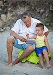 Grandfather and grandson spending time on the beach. Man watching the boy using tablet computer, they sitting on green blanket