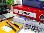 Red Ring Binder with Inscription Budget on Background of Working Table with Office Supplies, Laptop, Reports. Toned Illustration. Business Concept on Blurred Background.