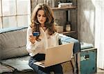 Elegant dressed young woman is sitting on couch and typing info from credit card to laptop