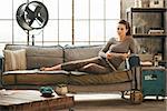 Thoughtful stylish brown-haired woman is sitting on couch and holding tablet PC in loft apartment