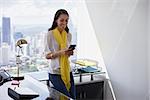 Young multiethnic Chinese Hispanic woman leaning on table in a modern office building, with sight of the city in background. She holds a mobile phone and smiles happy