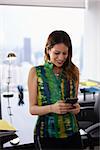 Young hispanic business woman leaning on table in a modern office building, with a beautiful sight of the city in background. She holds a mobile phone and smiles happy