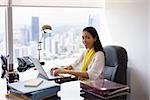 Portrait of mixed race business woman sitting in modern office in skyscraper. The secretary writes on laptop pc and smiles at camera. Medium shot