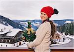 Mountain cottage hideaway brings calm and warmth into the winter season. Portrait of smiling young woman with Christmas tree standing on balcony overlooking the snow-capped mountains