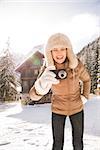 Christmas season in relaxed style of contemporary countryside living. Happy young woman standing in the front of a cosy mountain house and taking photos with digital camera