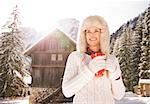 Christmas season in relaxed style of contemporary countryside living. Smiling young woman in white sweater holding red cup and looking into distance while standing in the front of cosy mountain house.