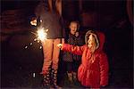 Side view of girl wearing red fur trim coat holding sparkler being ignighted by mother, smiling