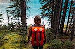 High angle rear view of mid adult woman carrying orange colour backpack standing in forest looking at water, Moraine lake, Banff National Park, Alberta Canada