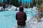 Rear view of mid adult man wearing hooded waterproof coat crouching by waters edge, Moraine lake, Banff National Park, Alberta Canada