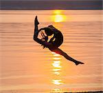 Side view of girl in silhouette by ocean at sunset in mid air, legs apart throwing head back