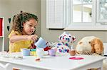 Girl in playroom sitting at table serving tea from toy tea set to soft toys
