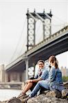 Two female tourists sitting on rock by river with Brooklyn Bridge in background