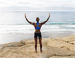 Young woman standing on rocks beside sea, arms raised, rear view