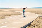 Mid adult woman cycling along pathway at beach, arms in air