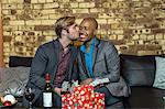 Male couple sitting on sofa, exchanging Christmas gifts, young man kissing partner on cheek