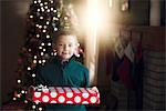 Boy in front of christmas tree holding gift looking at camera smiling