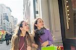 Young female adult twins with shopping bags looking up at shop window