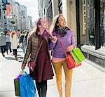 Young female adult twins carrying shopping bags strolling and chatting on city sidewalk