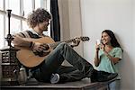 Loft living. A young man playing guitar to an appreciative audience, a young woman holding a smart phone and taking a photograph.