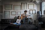 Loft decor. A wall hung with pictures in frames, reversed to show the backs. A man playing a guitar and a woman sitting on a sofa.