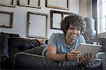 Loft decor. A wall hung with pictures in frames, reversed to show the backs. A man lounging on a sofa, using a digital tablet.