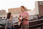 A man playing a guitar to a woman, on a rooftop terrace overlooking a city at dusk.