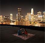 A couple sitting on a rug on a rooftop overlooking a city lit up at night.