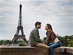 A couple seated facing each other on a wall in the city of Paris with the Eiffel Tower in the background.