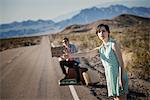 A young couple, man and woman, on a tarmac road in the desert hitchiking, with a sign saying Vegas or Bust.