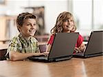 Two children seated at desks in class using laptop computers.