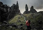 Two people with rucksacks on a narrow path rising to a dramatic landscape of rock pinnacles on the skyline towering above them, under an overcast sky with low cloud.