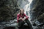 A man sitting on a rock by a waterfall, holding a flask. Winter hiking.