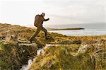 A man with a rucksack and winter clothing leaping across a small stream in an open exposed landscape.