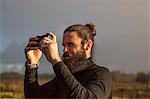 A man standing using a smart phone to take a photograph under a grey sky.