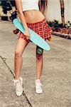 Close up of a sporty lady in a sexy red tartan mini skirt holds her blue penny skateboard shortboard while stands on the tropical street. Outdoor lifestyle picture on a sunny summer day.