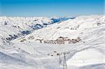 View down a snowy ski piste in alpine mountain valley with cable car lift and village