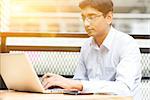 Candid Asian Indian businessman using laptop computer, modern building with golden sunlight at background.