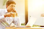 Man using laptop computer while drinking a cup hot milk tea, outdoor cafe, beautiful sunlight during sunset.