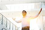 Asian Indian businessman arms outstretched celebrating success, modern office building with morning sunlight as background.