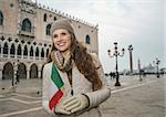 Delightful Venice, Italy can help make the most of your next winter getaway. Happy young woman tourist with Italian flag standing on St. Mark's Square near Dogi Palace and looking into the distance