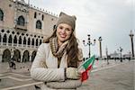 Venice, Italy can help make the most of your next winter getaway. Portrait of smiling young woman tourist with Italian flag standing on St. Mark's Square near Dogi Palace and looking into the distance
