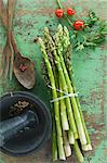 A bundle of fresh green asparagus, tomatoes and parsley on a wooden table, peppercorns in a mortar and salad servers