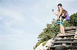 Low angle side view of young man standing on wooden steps using smartphone to take photograph, Cala Luna, Sardinia, Italy