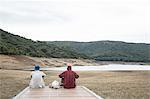 Rear view of young men sitting on wooden pier looking at mountain range, Nuoro, Sardinia, Italy