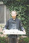 Front view of bearded mid adult man holding tray with clay pot looking at camera smiling