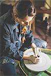 Young woman sitting at pottery wheel shaping clay, looking down