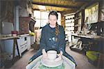 Front view of young woman in workshop sitting at pottery wheel making clay pot, looking down