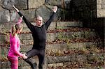 Mid adult man on stone steps arms raised standing on one leg, young woman helping