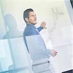 Business man making a presentation in front of whiteboard. Business executive delivering a presentation to his colleagues during meeting or in-house business training. View through glass.