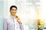 Portrait of Asian Indian business man smiling, leaning on modern office building block, beautiful golden sunlight at background.